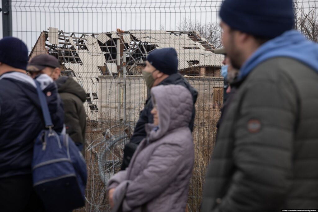 Donbas Frontliner / КПВВ Станиця Луганська. Люди стоять у черзі до модуля прикордонників на тлі зруйнованих у 2014-2015 роках будинків